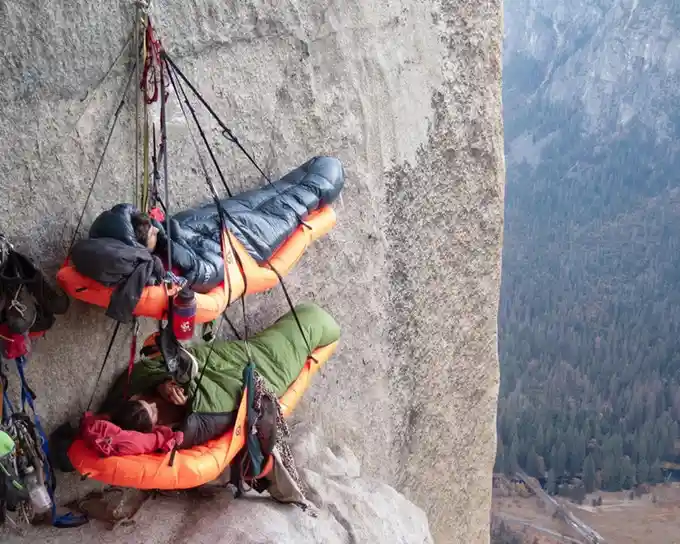 Image of two mountain climbers resting in climber's bivouacs. They look like intricately rigged contraptions of ropes and climbing equipment suspended off the side of a cliff. The climbers look very cozy in sleeping bags inside the contraptions.